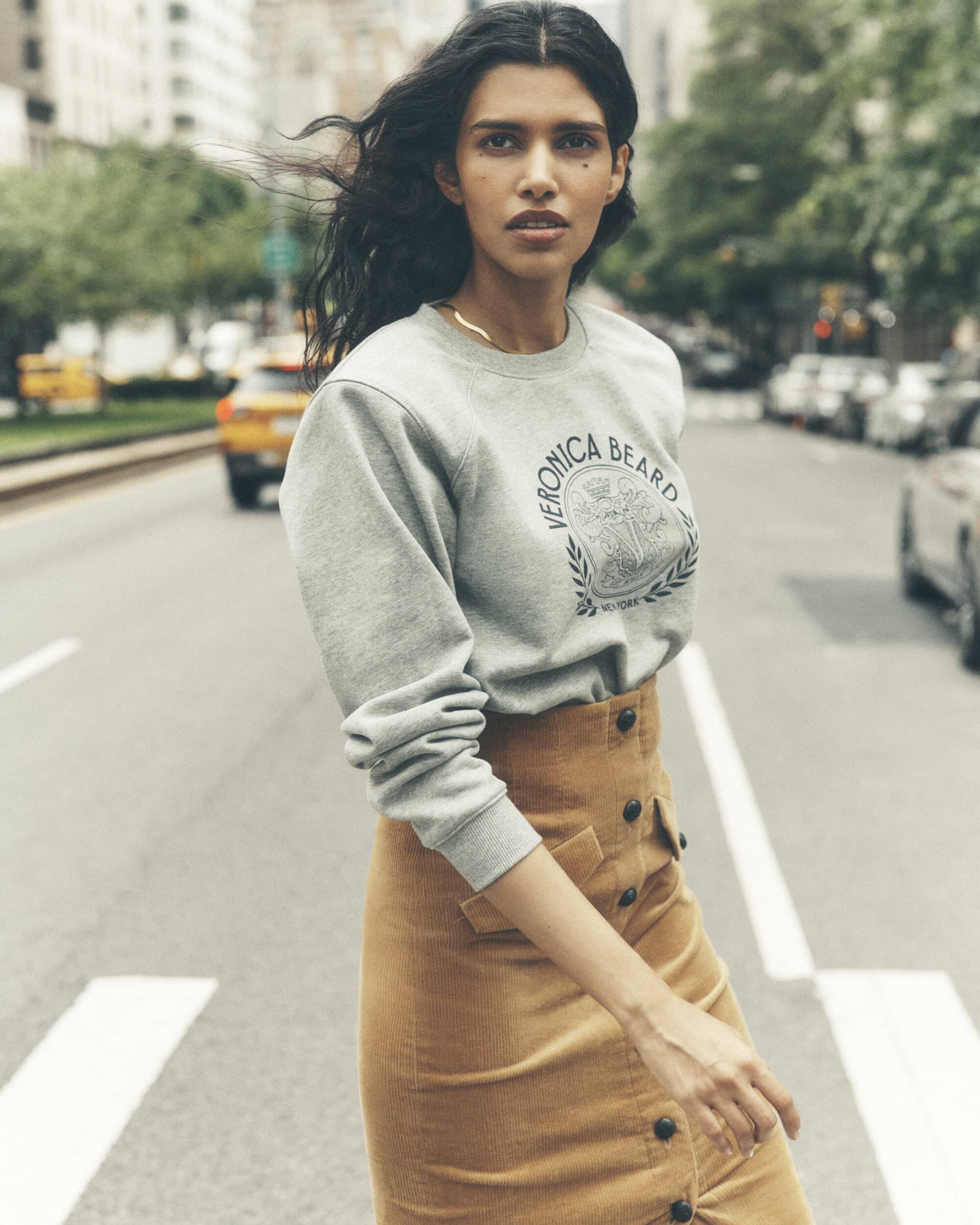 Woman walks on a crosswalk in New York City, wearing a grey Veronica Beard sweatshirt and a brown corduroy skirt with buttons.
