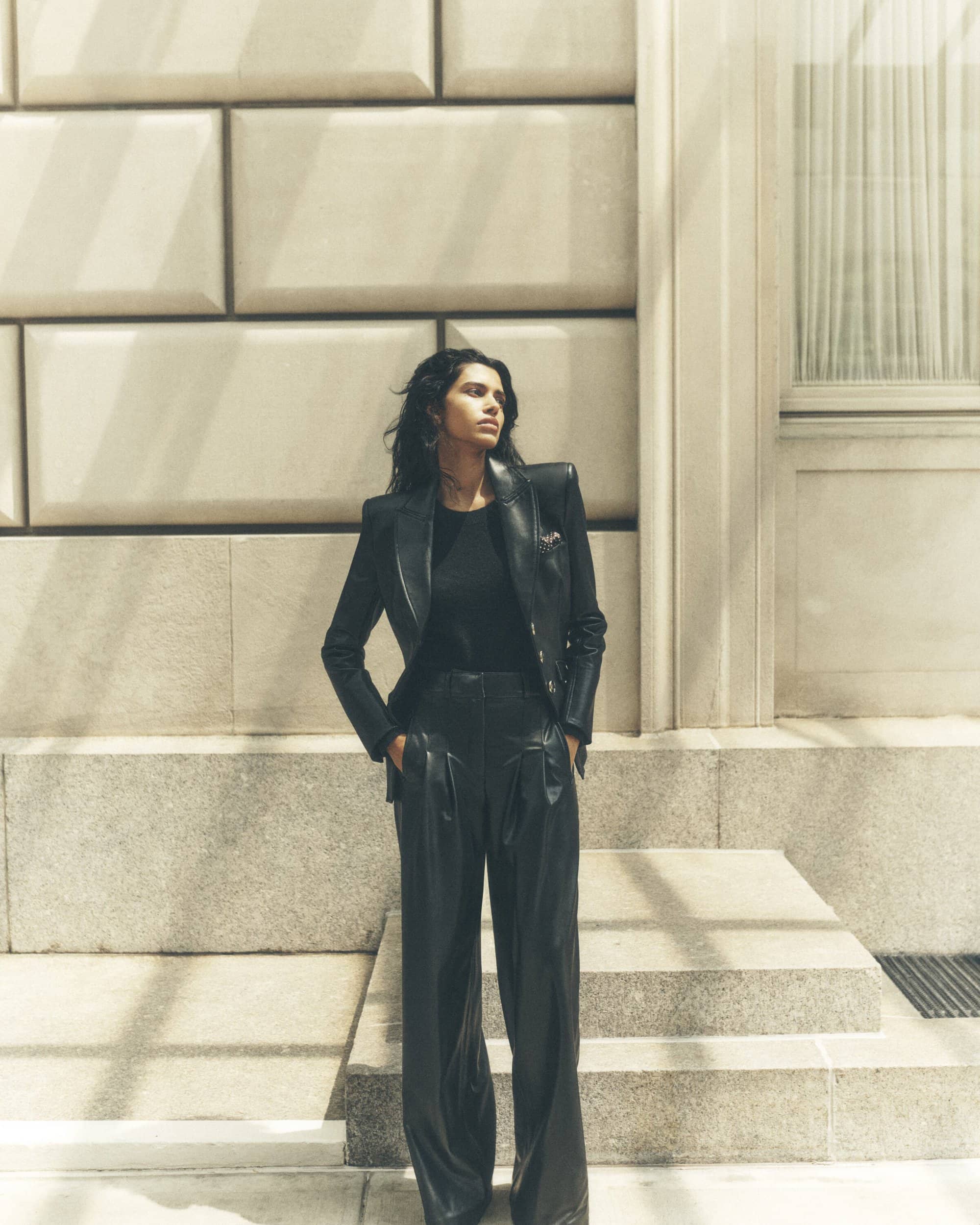 A stylish woman in a black leather jacket and pants stands confidently against a textured stone wall, bathed in soft, diffused light.