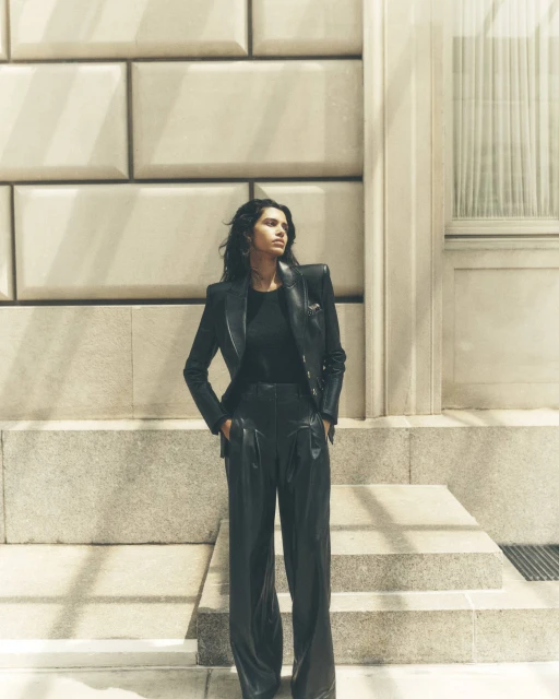 A stylish woman in a black leather jacket and pants stands confidently against a textured stone wall, bathed in soft, diffused light.
