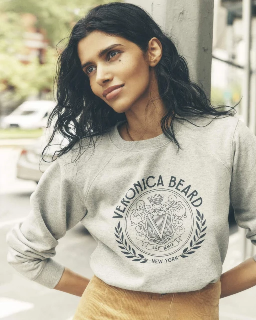 Woman wearing a Veronica Beard logo sweatshirt and mustard skirt. The background is a city street. The shot is medium and eye-level.