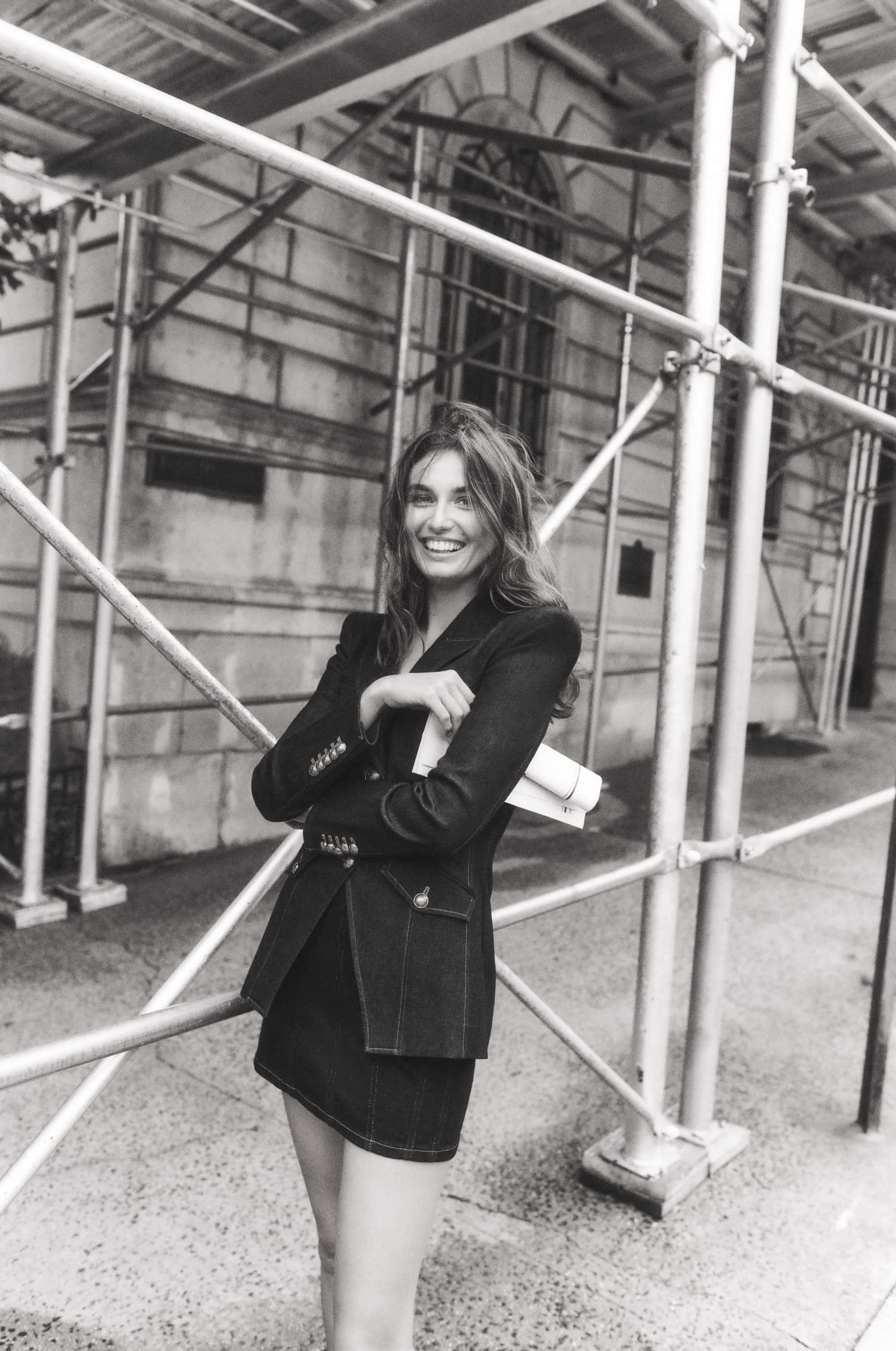 Black and white portrait of a smiling woman in a blazer and skirt standing near scaffolding, holding papers, in an urban environment.