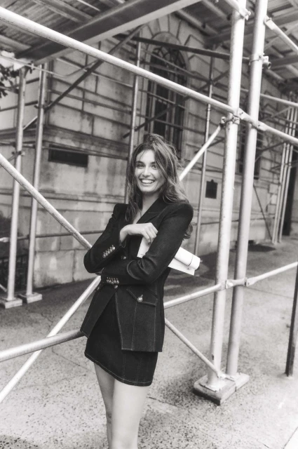 Black and white portrait of a smiling woman in a blazer and skirt standing near scaffolding, holding papers, in an urban environment.