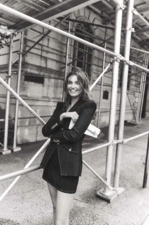Black and white portrait of a smiling woman in a blazer and skirt standing near scaffolding, holding papers, in an urban environment.