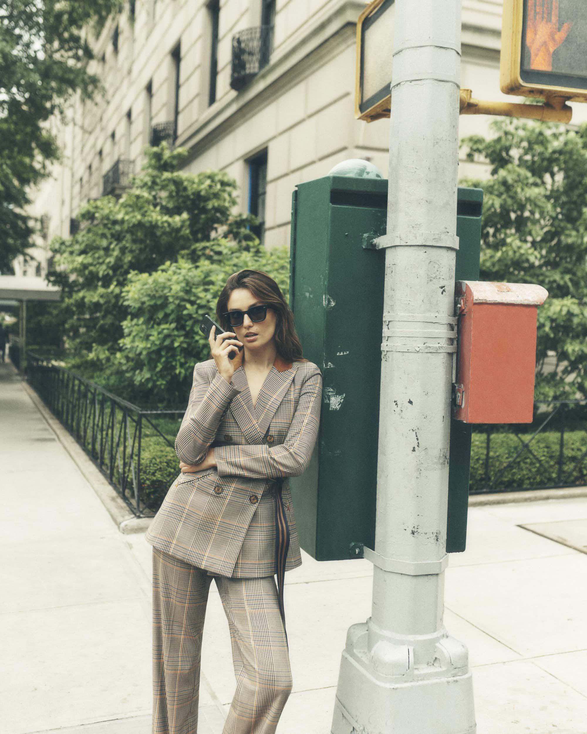 Woman in a plaid suit uses a smartphone on a city sidewalk. The woman stands near a green utility box and crosswalk sign post.