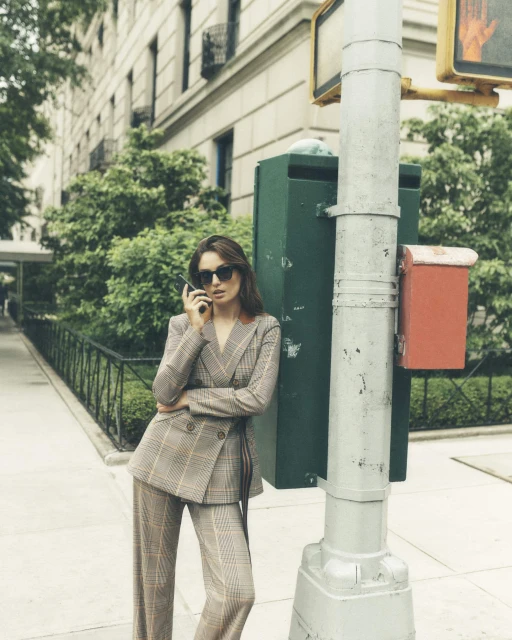 Woman in a plaid suit uses a smartphone on a city sidewalk. The woman stands near a green utility box and crosswalk sign post.
