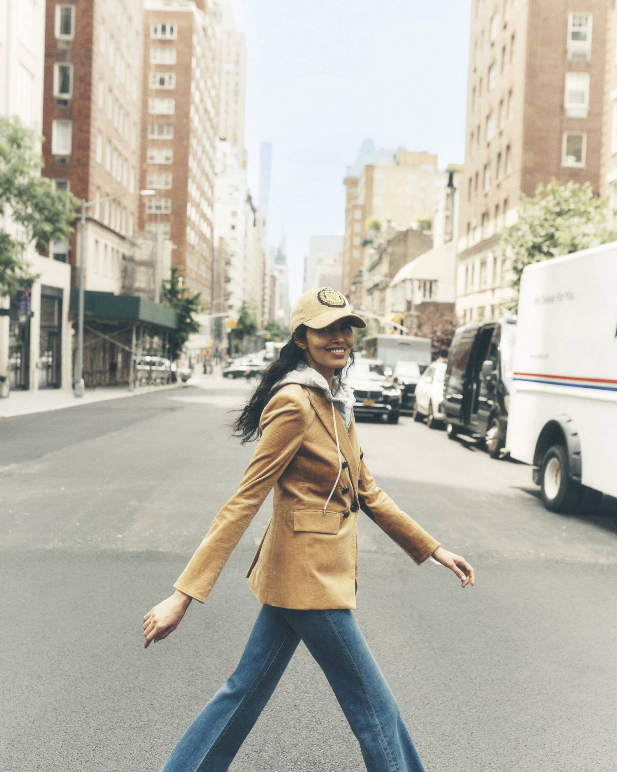 A young woman confidently crosses a New York City street in a tan blazer and jeans, with buildings and vehicles forming an urban backdrop.