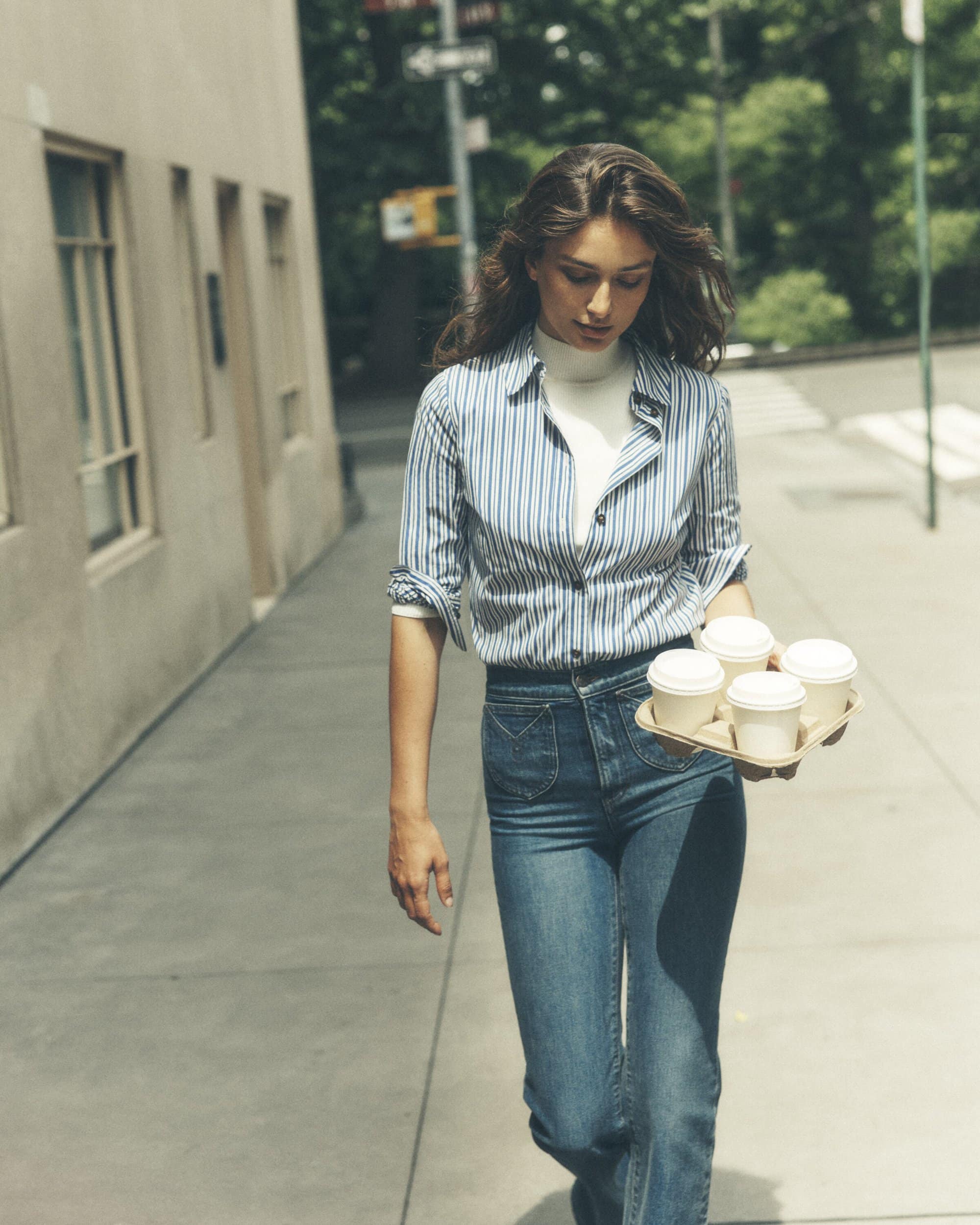 A young adult woman walks along a city street carrying a tray of coffee cups on a sunny day with buildings in the background.
