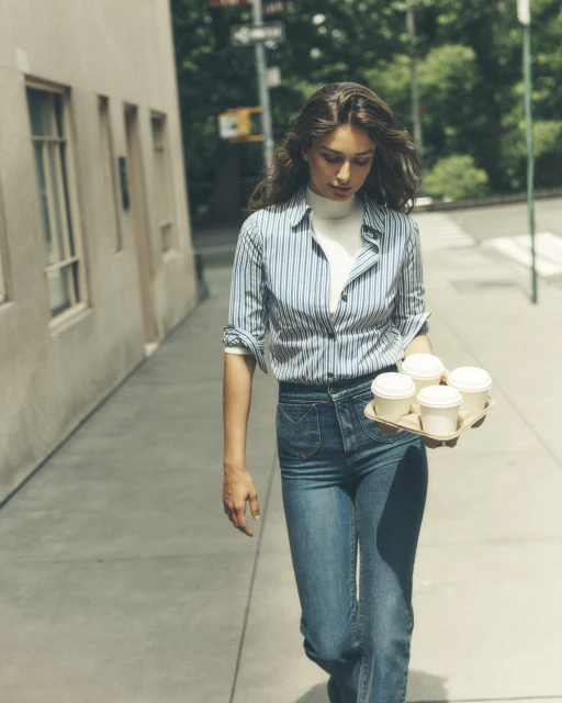 A young adult woman walks along a city street carrying a tray of coffee cups on a sunny day with buildings in the background.