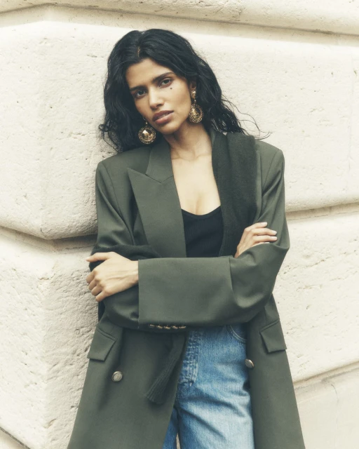 A woman in an olive green blazer stands indoors against a textured wall, posing with her arms crossed, wearing statement earrings.