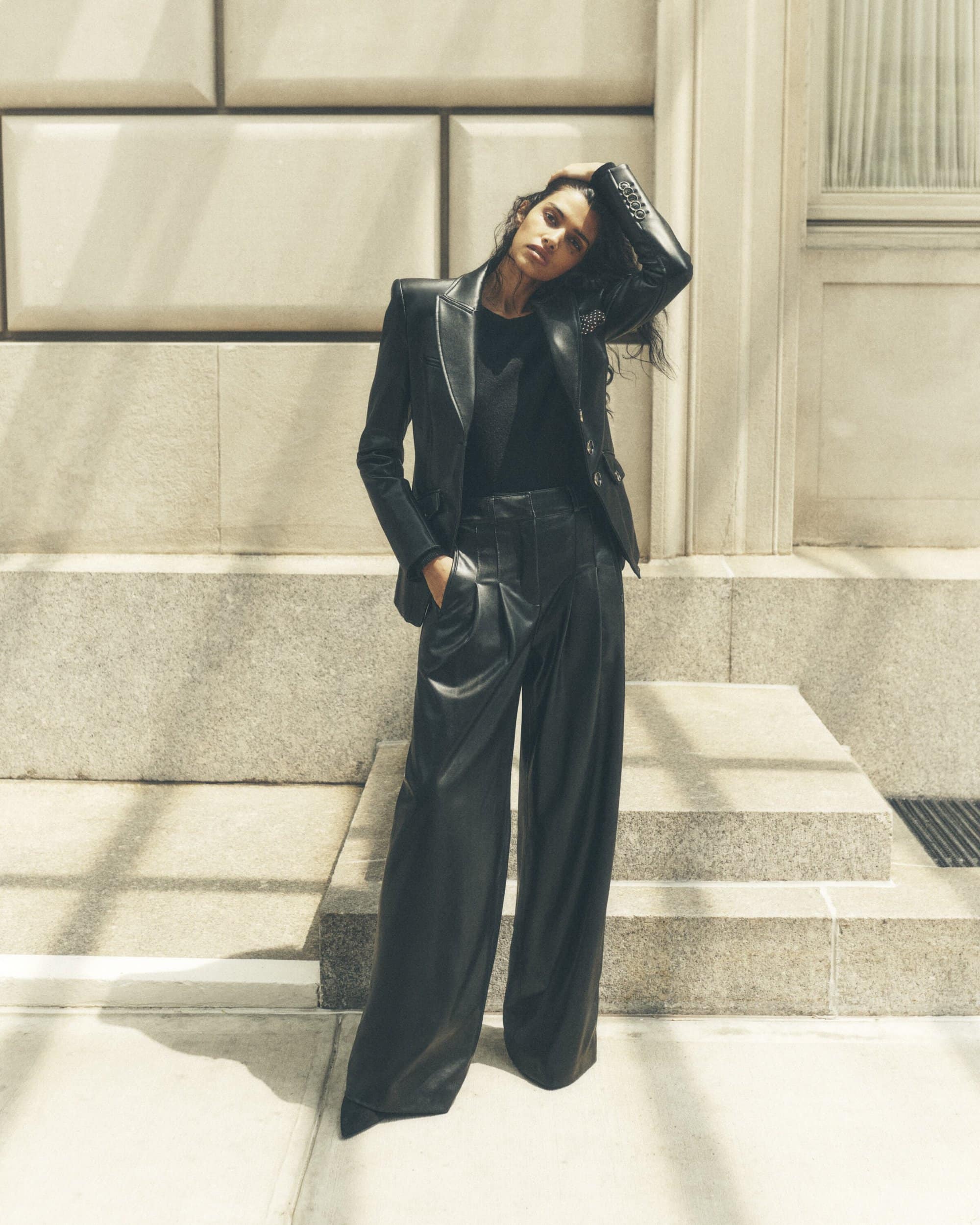 Brunette woman in a black leather blazer and matching wide-leg pants poses against an architectural background in soft, natural light.
