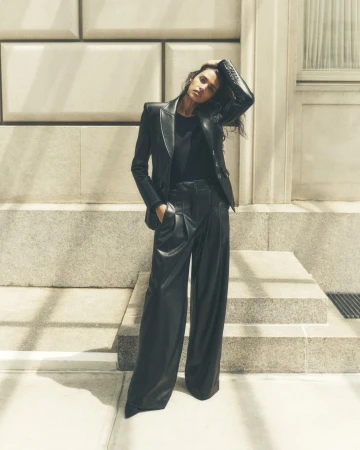 Brunette woman in a black leather blazer and matching wide-leg pants poses against an architectural background in soft, natural light.