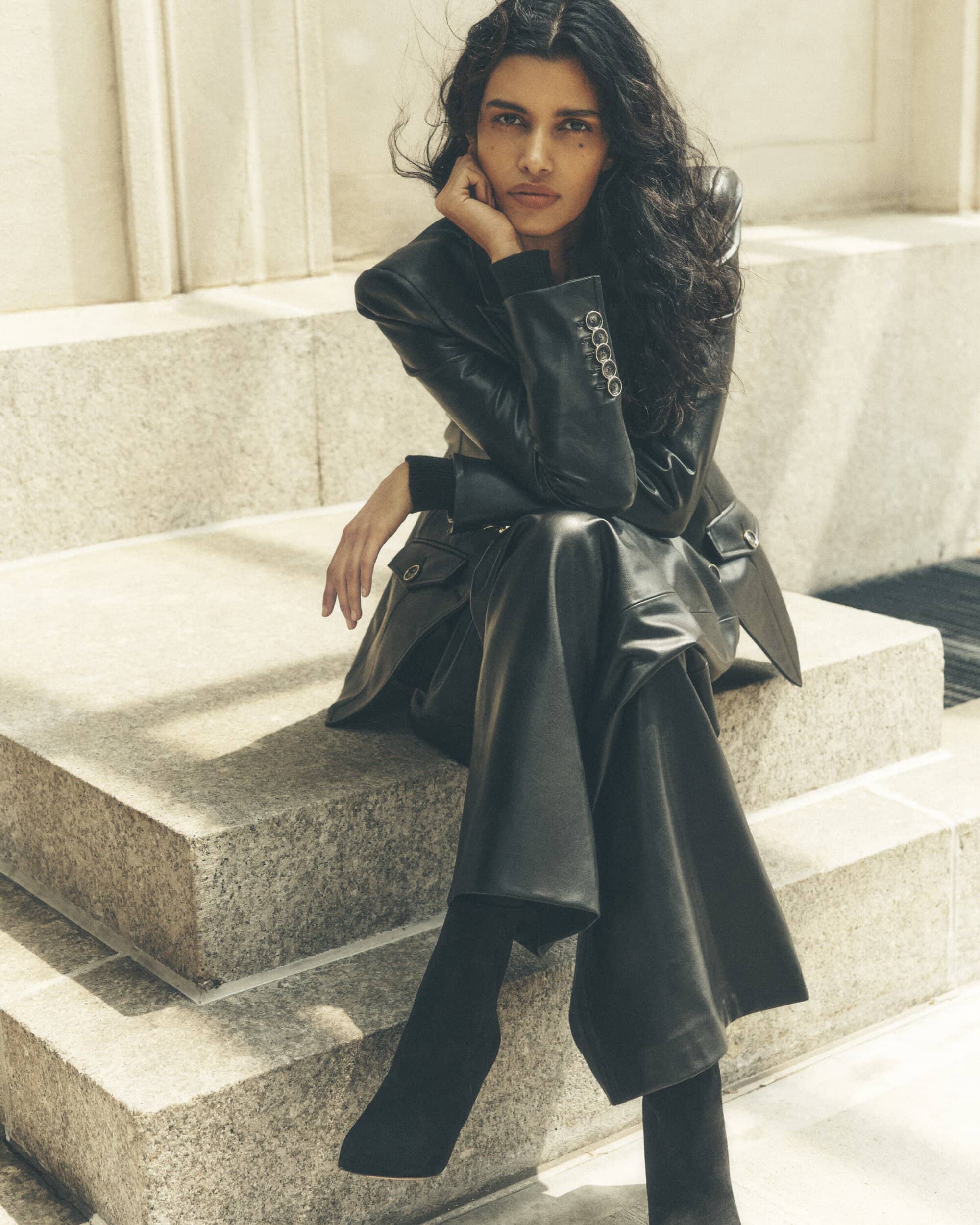 Brunette woman in a black leather jacket and wide-leg pants poses on stone steps in an outdoor setting, looking at the camera.