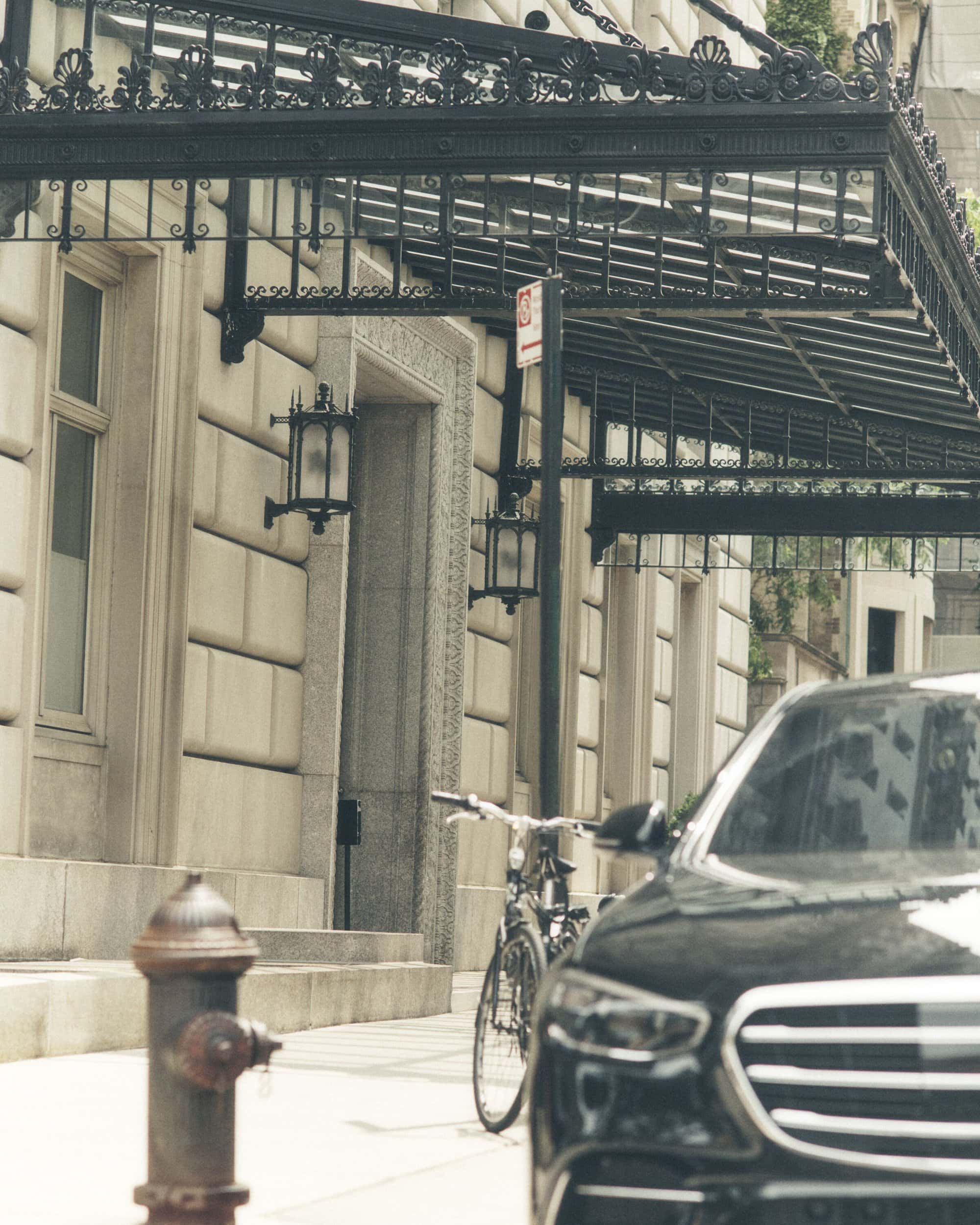 Exterior shot of a luxury car parked on a city street with a vintage bicycle and fire hydrant, building with awning in background.