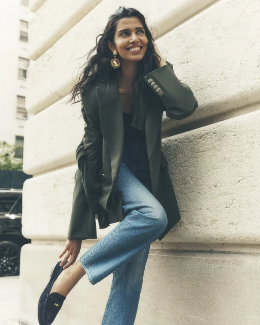 A smiling woman in an olive-green blazer and jeans poses against an urban building wall, looking up and to the right, stylish and happy.