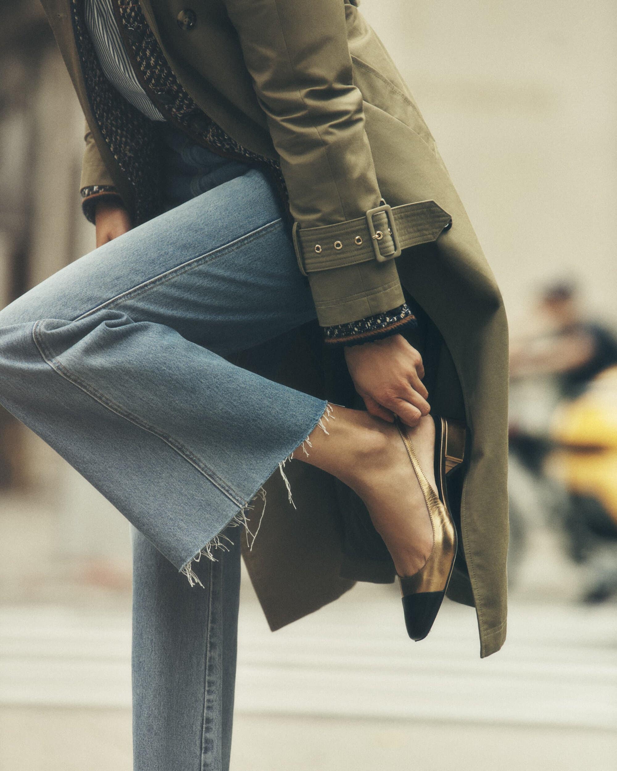 Woman in a trench coat adjusts her gold and black heeled shoe on a city street, with blurred figures in the background.