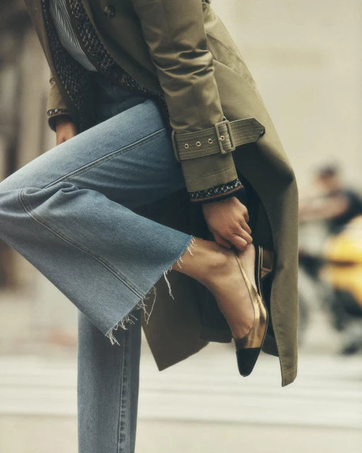 Woman in a trench coat adjusts her gold and black heeled shoe on a city street, with blurred figures in the background.