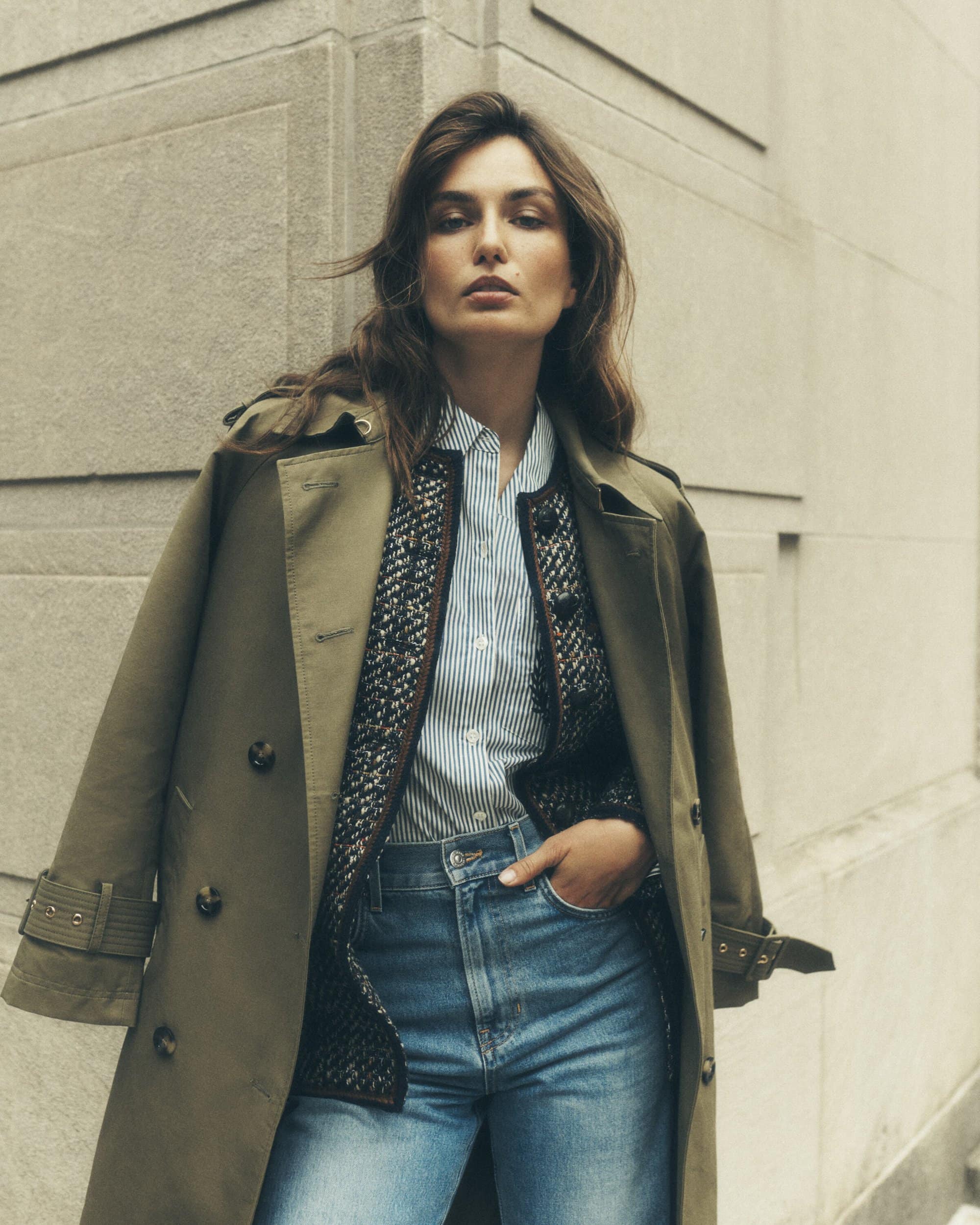 Woman in an olive trench coat and blue jeans posing against an exterior wall in a medium shot, fashion editorial, neutral color grading.