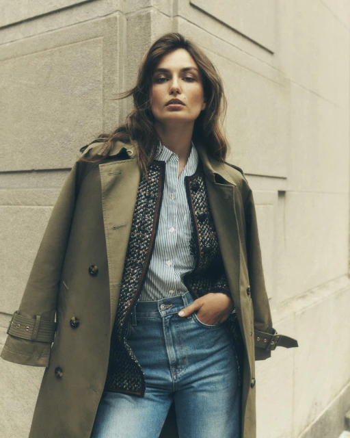 Woman in an olive trench coat and blue jeans posing against an exterior wall in a medium shot, fashion editorial, neutral color grading.