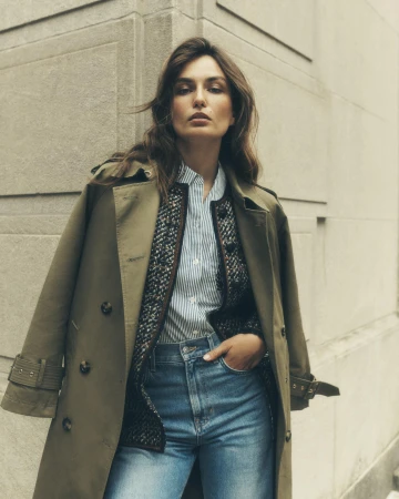 Woman in an olive trench coat and blue jeans posing against an exterior wall in a medium shot, fashion editorial, neutral color grading.
