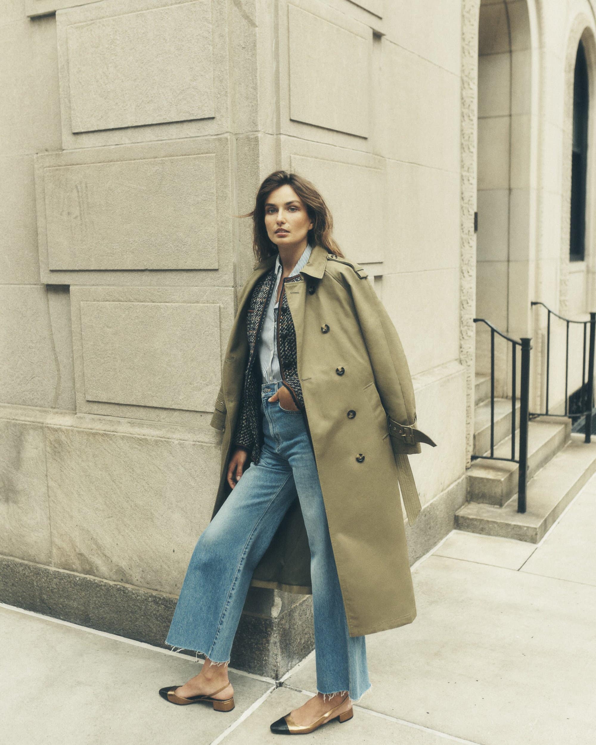 A woman in a tan trench coat and light-wash jeans poses on a city street with a building and wrought-iron fence in the background.