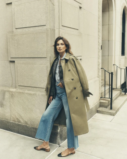 A woman in a tan trench coat and light-wash jeans poses on a city street with a building and wrought-iron fence in the background.
