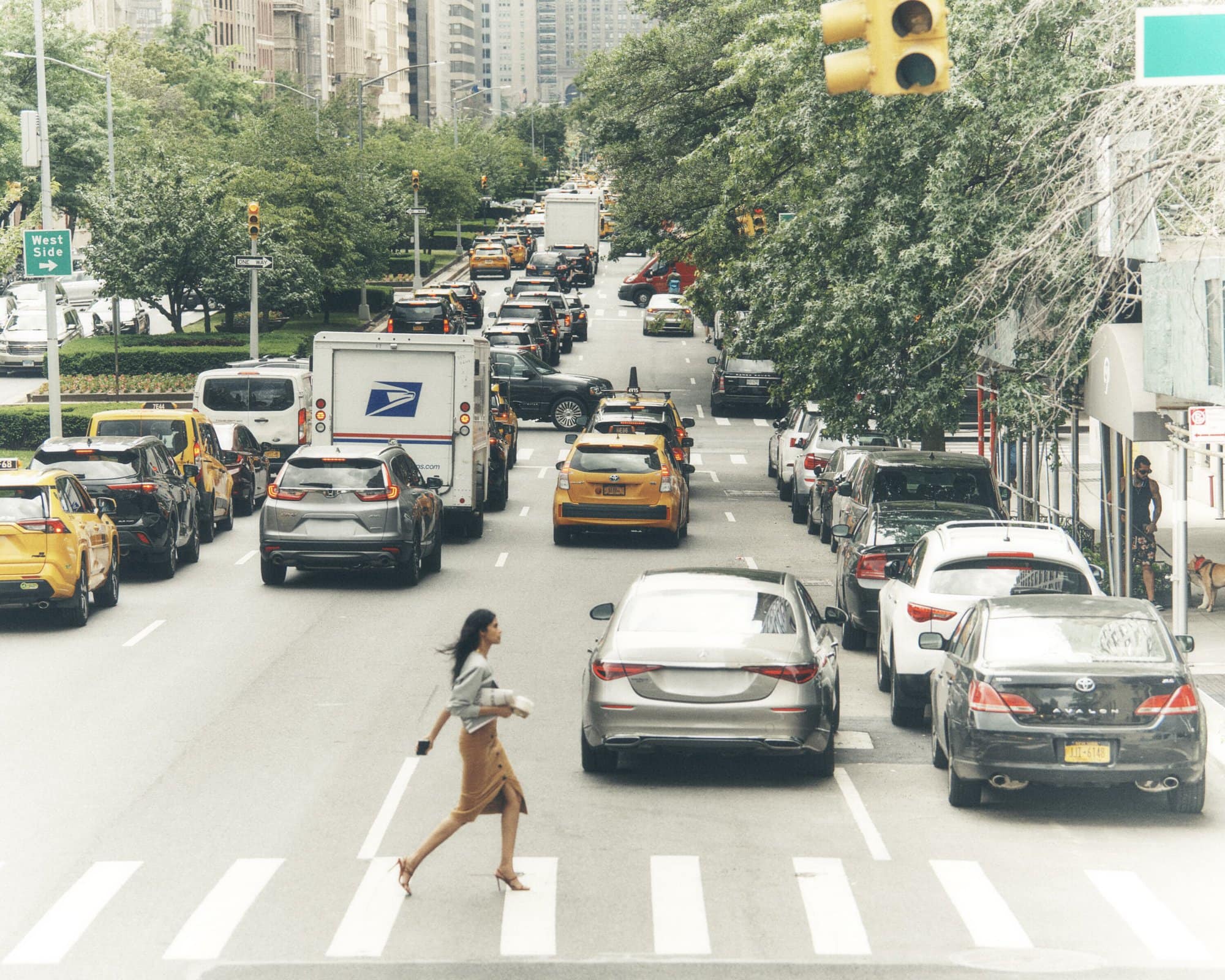Woman in a tan skirt walks across a city street at a crosswalk with traffic and skyscrapers in the background on an overcast day.