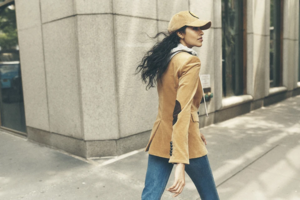 Woman in tan blazer, blue jeans, and cap walking in city; wind in her hair, side profile, architectural details in background, outdoor setting.