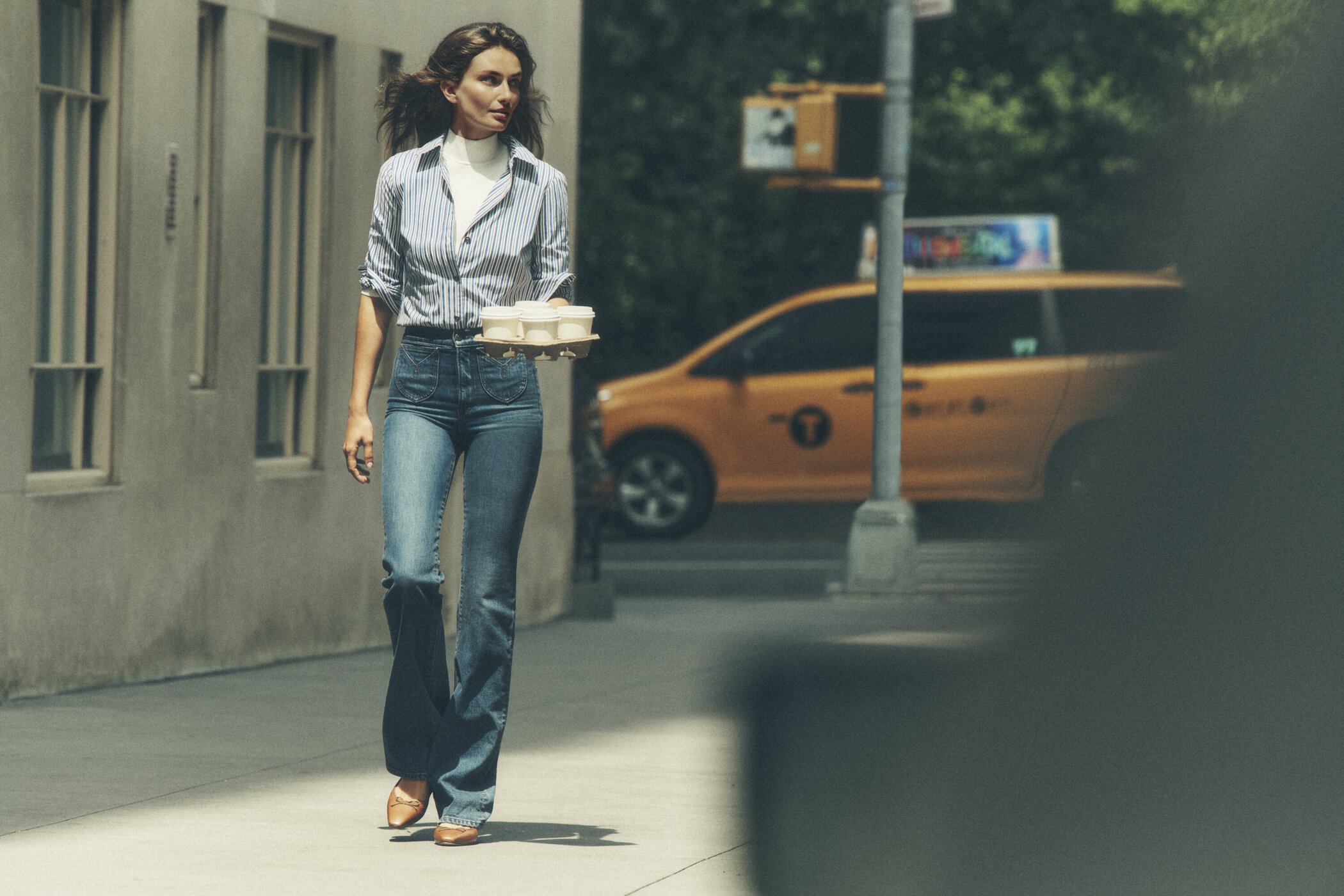 Woman walks on a city sidewalk carrying a tray of containers, wearing a striped shirt, jeans, and heels, with a taxi in the background.