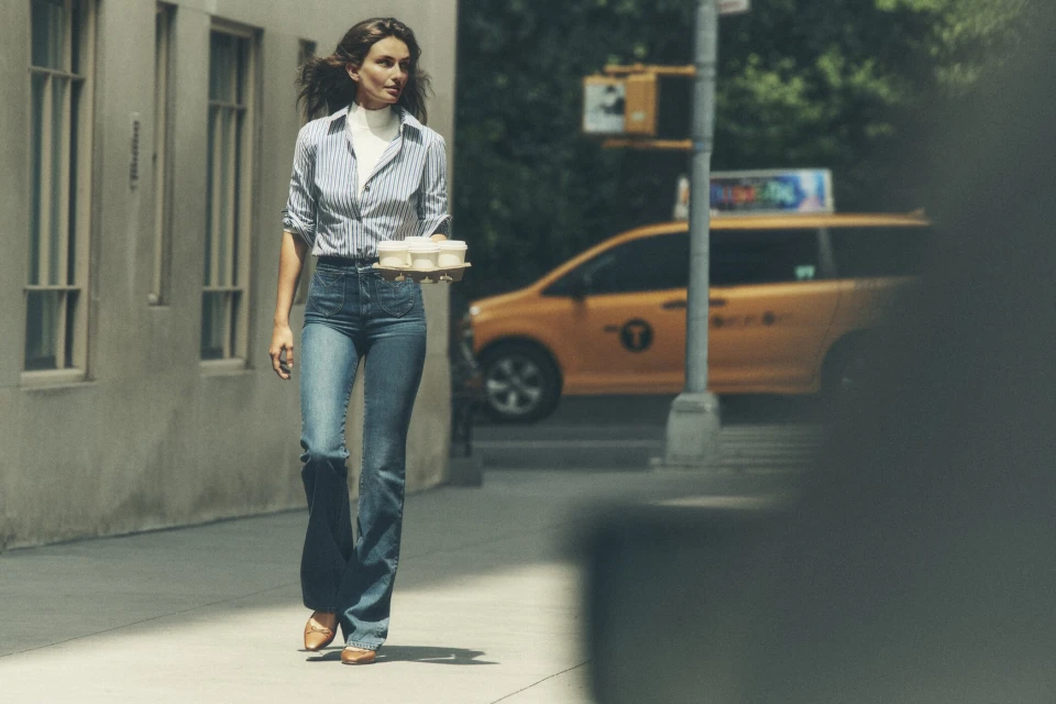 Woman walks on a city sidewalk carrying a tray of containers, wearing a striped shirt, jeans, and heels, with a taxi in the background.