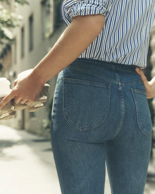 Back view of a woman in jeans holding coffee cups on a city street with buildings in the background, capturing an urban lifestyle moment.