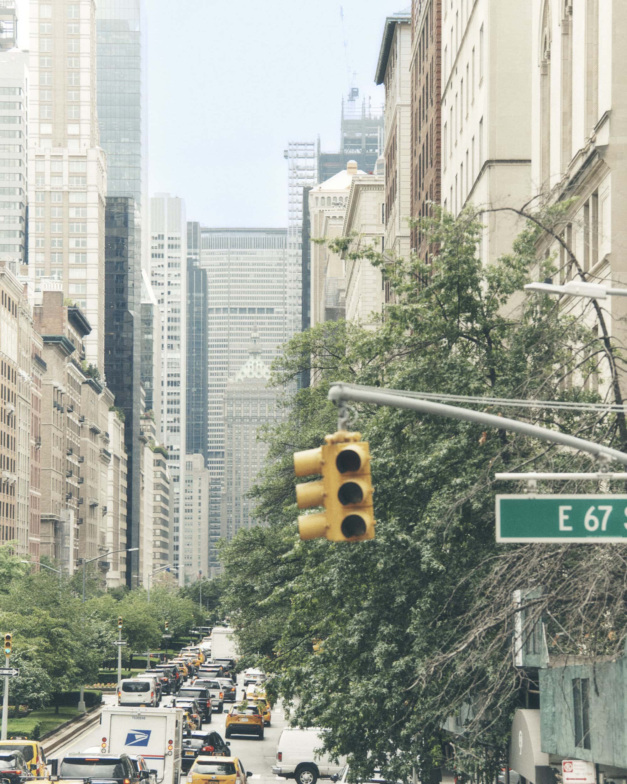 Street view in New York City, featuring a traffic light, cars, buses, and tall buildings on a cloudy day. Street sign visible.