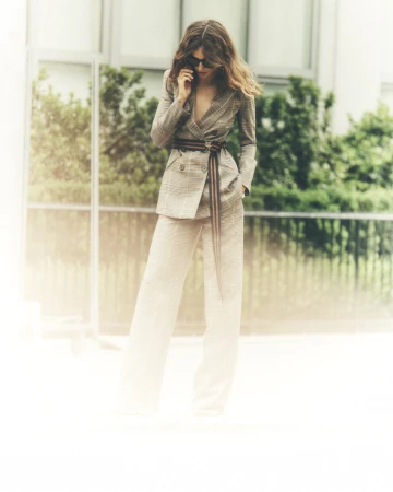 Woman in a plaid suit wearing sunglasses stands outdoors with green foliage. She has a belt and is looking down.