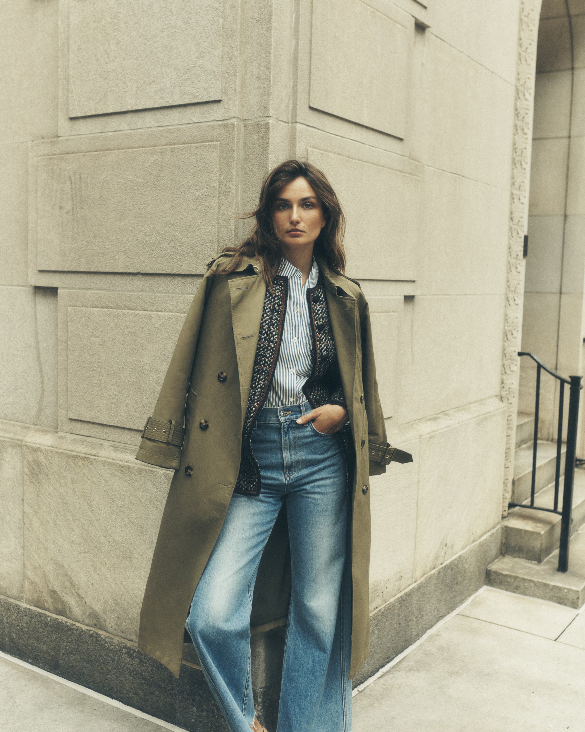 Woman posing in a trench coat and jeans in an urban setting, leaning against a building. Fashion photography with a neutral color palette.