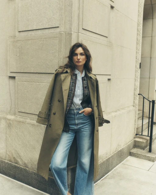 Woman posing in a trench coat and jeans in an urban setting, leaning against a building. Fashion photography with a neutral color palette.