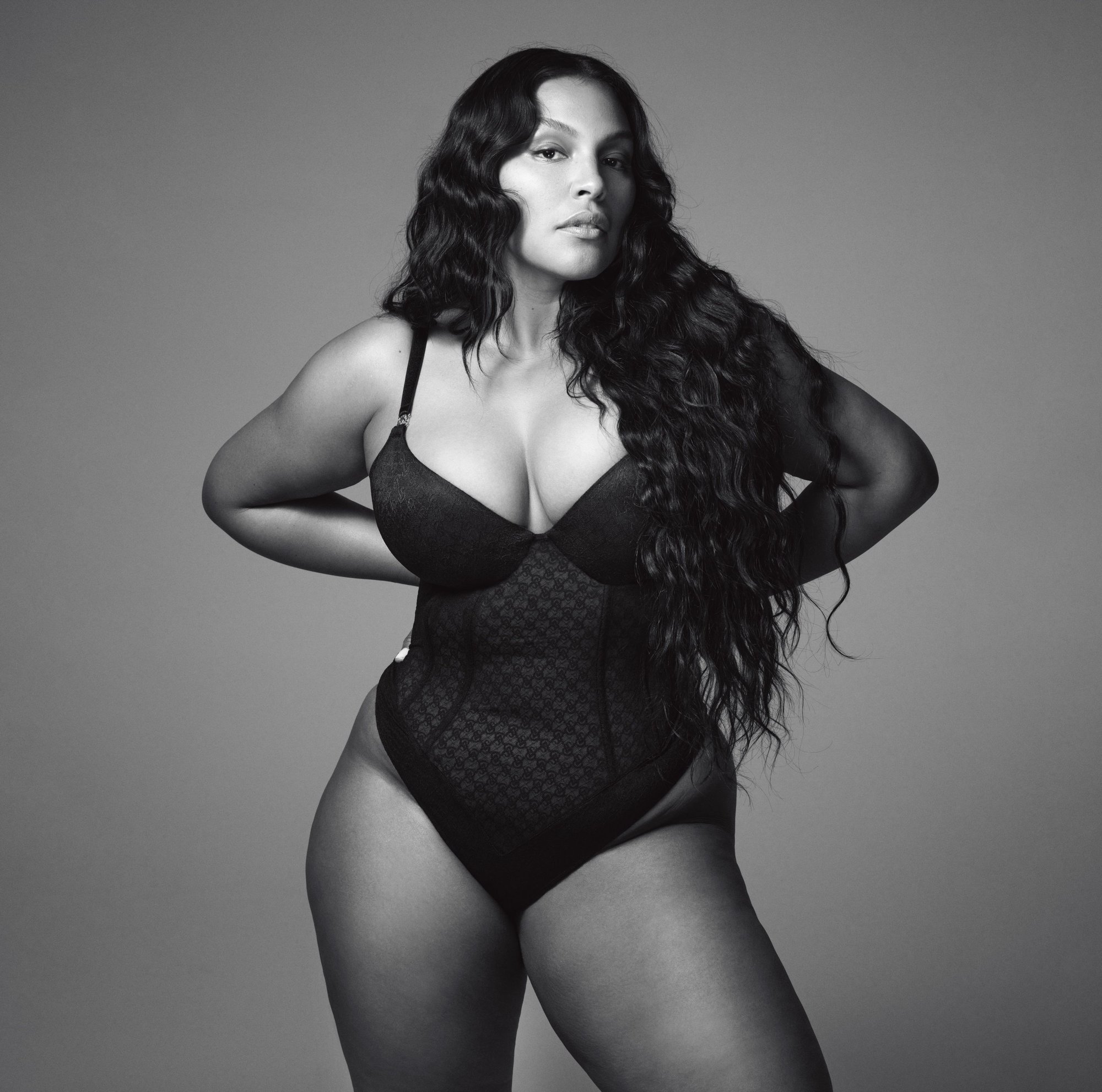 Portrait of an adult woman with long dark wavy hair in a black lacy bodysuit against a grey background, fashion and beauty shot.