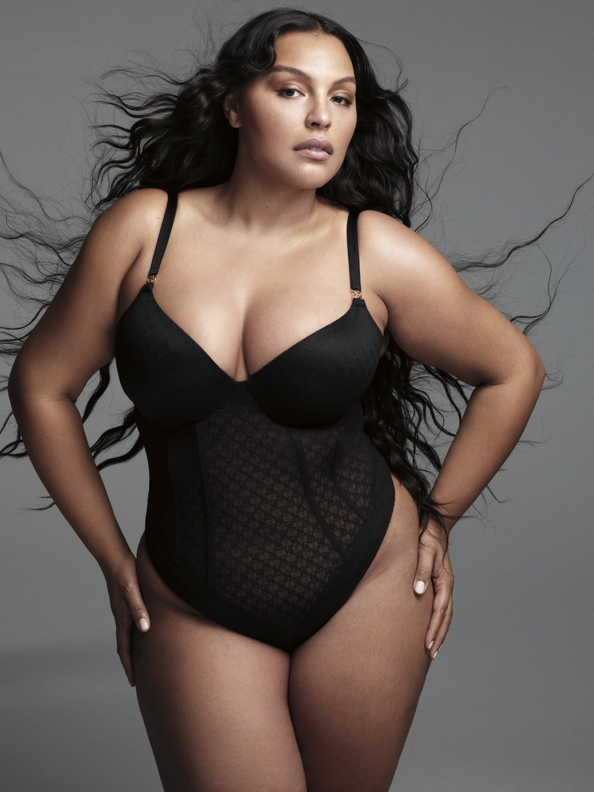 Portrait of plus-size adult woman in black bodysuit. She has wavy dark hair and light skin. Studio shot with gray background.