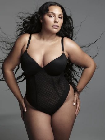Portrait of plus-size adult woman in black bodysuit. She has wavy dark hair and light skin. Studio shot with gray background.