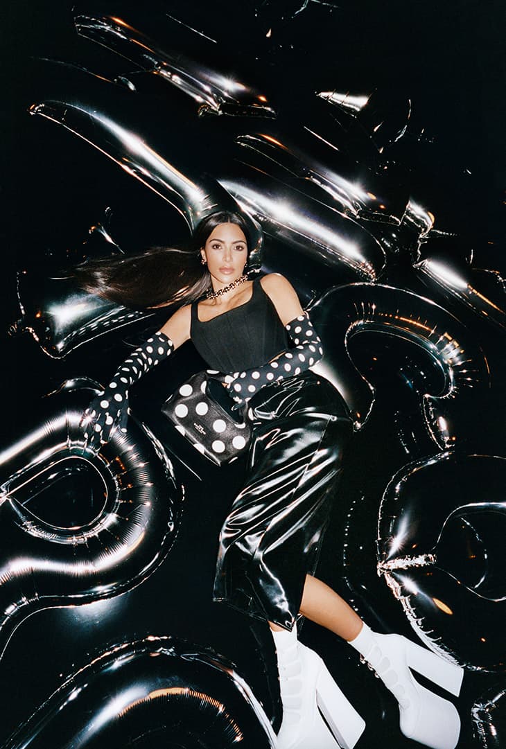 Woman lying on a black surface surrounded by silver letter-shaped balloons, wearing a black and white polka dot patterned outfit.