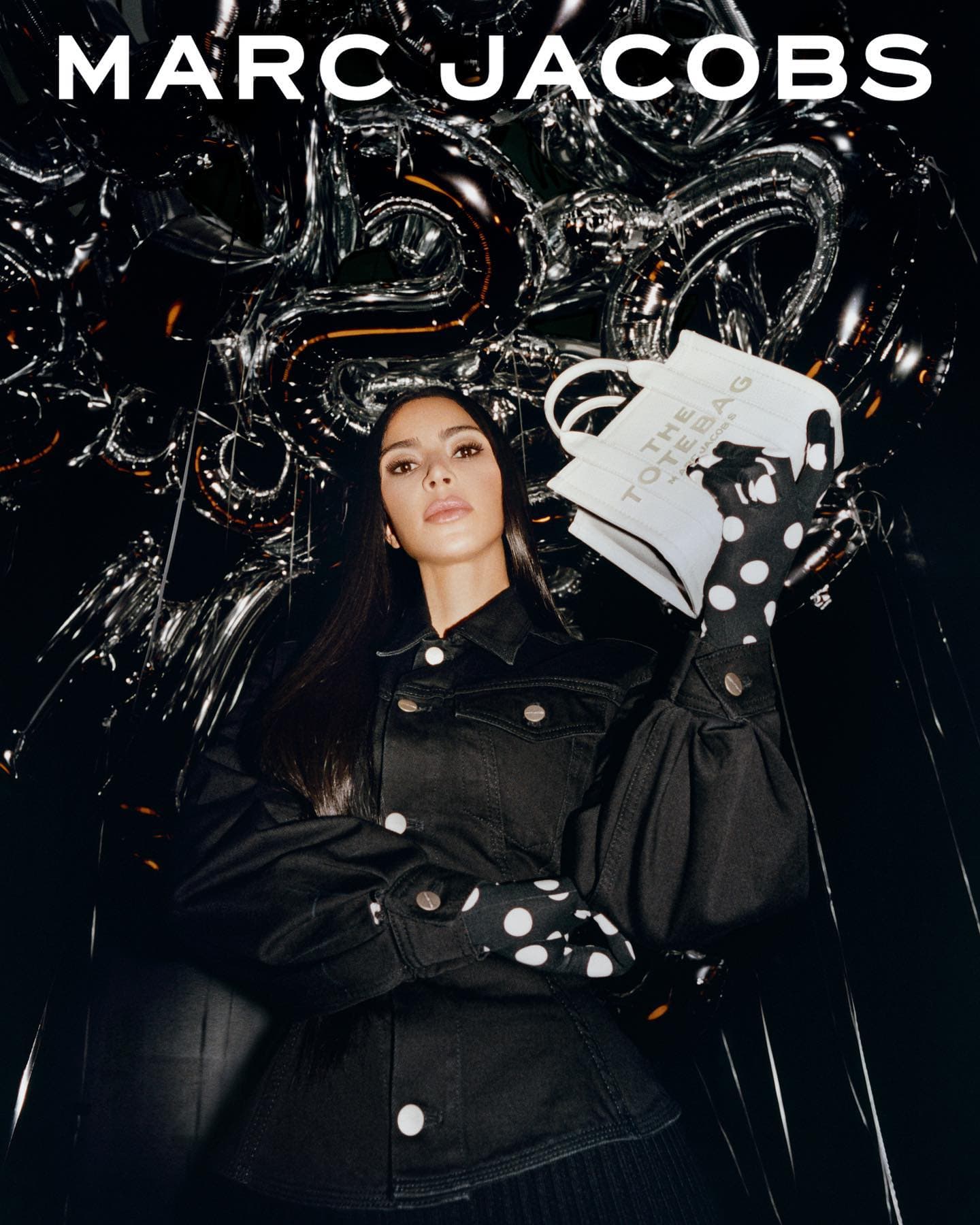 Fashion shot features a woman in a black denim jacket holding a Marc Jacobs tote bag, posing against a reflective balloon backdrop.