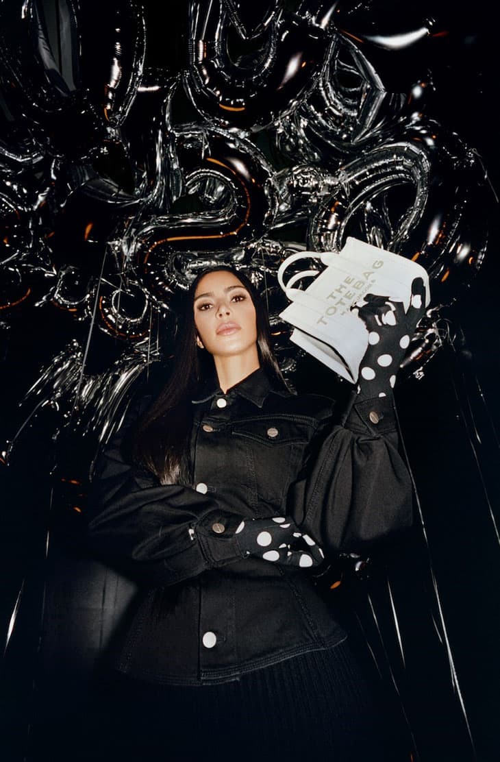 Woman poses with a white tote bag and silver balloon decor. She wears a black denim jacket and polka dot gloves in a photo studio.