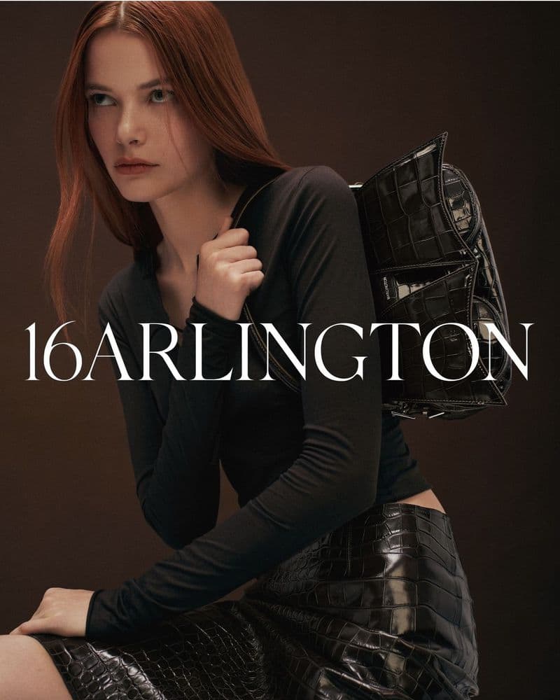 A woman with auburn hair poses with a black crocodile-skin purse and skirt against a solid brown backdrop for a fashion editorial.
