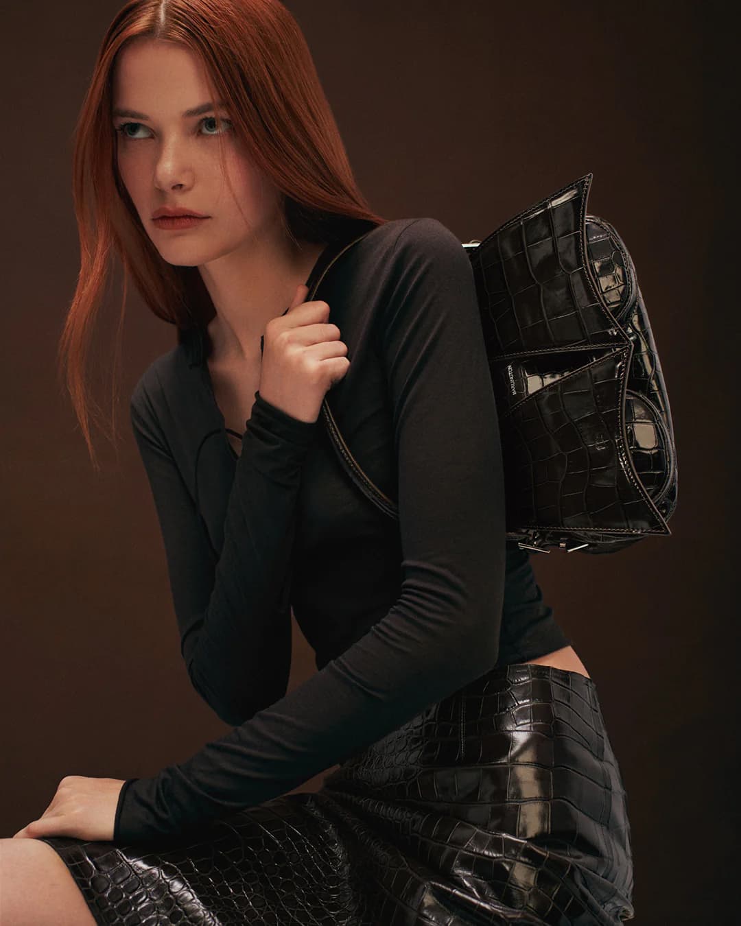 Fashion shot of a redhead woman in black long-sleeve top and reptile-print skirt with a black reptile-print shoulder bag.