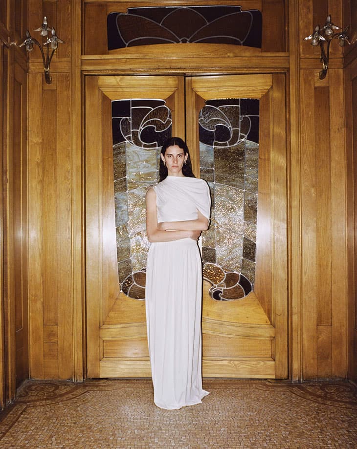 Elegant woman in white dress poses indoors in front of a wooden door with stained glass accents, in a full shot and eye-level view.