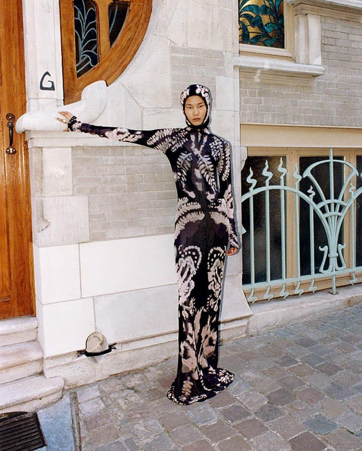 Adult female in hooded patterned dress poses beside a building with wooden door and pale brick and stone exterior.