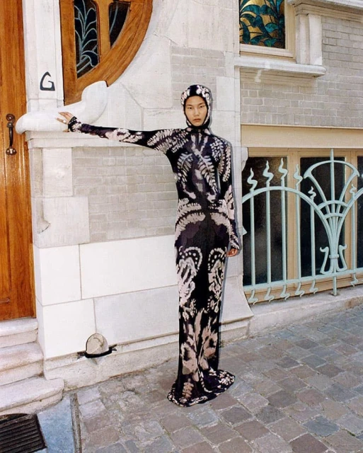 Adult female in hooded patterned dress poses beside a building with wooden door and pale brick and stone exterior.