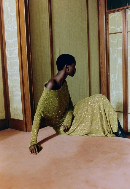 A Black woman sits in a green sequin dress with black boots on a carpet indoors, gazing to the right near a paneled wall.
