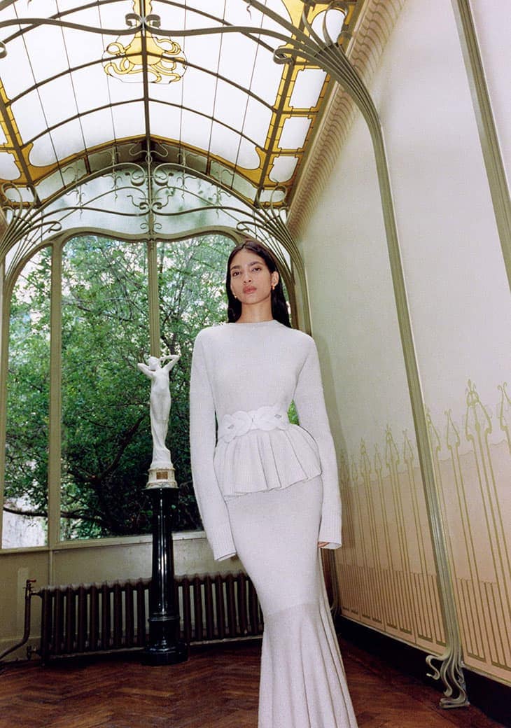 Elegant woman in white dress posing in a bright hallway with large windows, Art Deco style architecture, and natural outdoor light.