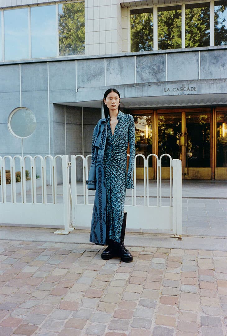 An Asian female fashion model poses outdoors in front of a building wearing a printed blue dress with black boots and a denim jacket.
