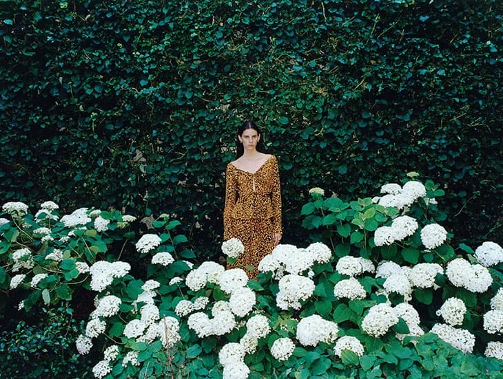 An adult woman with long brown hair in a patterned dress stands in a garden surrounded by white flowers and green foliage.