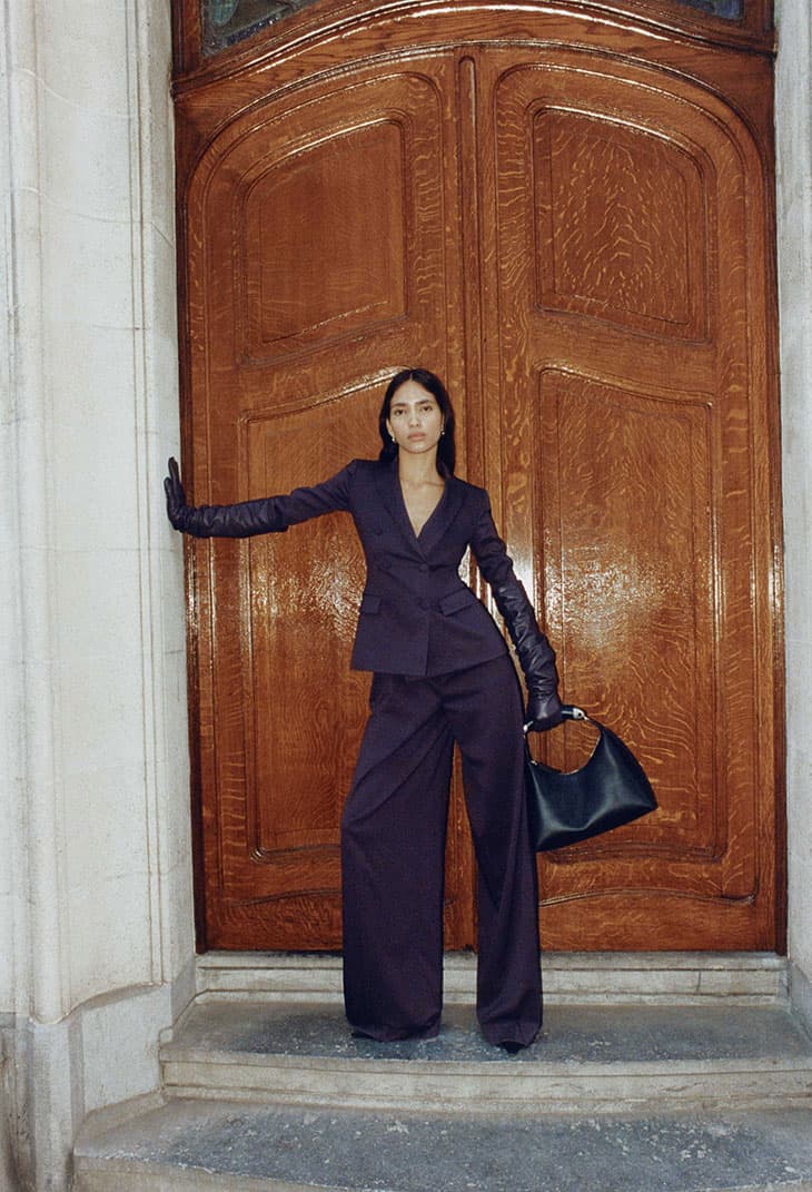 A stylish adult female in a purple suit and long gloves poses in front of a large wooden door, holding a black purse. Fashion.